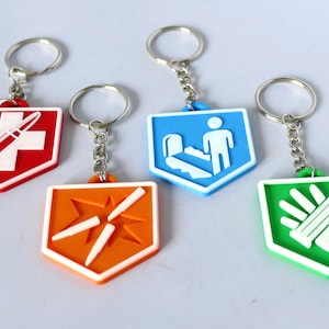 May include: Four shield-shaped keyrings with metal rings and chains. The red keyring features a white cross and bullet, the orange one shows an explosion, the blue one depicts a person on an escalator, and the green one displays a hand.