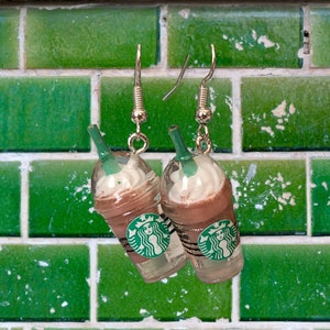 May include: A pair of silver dangle earrings featuring a green and brown Starbucks Frappuccino charm. The charms are shaped like a tall cup with a white whipped cream top and a green Starbucks logo.