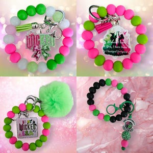 May include: Four beaded keychain bracelets in various color combinations. One features green, white, and pink beads with a green tassel and a charm that says "Wicked Era". Another has pink and green beads with a charm that says "Because I knew you, I have been changed for good".