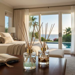 May include: Two clear glass reed diffusers with gold-toned collars and wooden reeds. The left bottle contains light green stones, while the right bottle contains multi-colored pebbles. Both are filled with liquid and sit on a dark wooden surface.