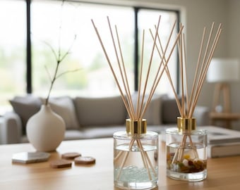 Op de afbeelding: Twee glazen geurstokjesdiffusers met goudkleurige hals en houten stokjes. De linker diffuser bevat doorzichtige glazen kralen, terwijl de rechter diffuser gevuld is met kleine, veelkleurige decoratieve steentjes in vloeistof.