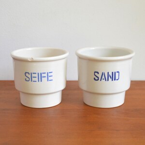Könnte beinhalten: Zwei weiße Keramikbehälter mit blauem Text. Einer trägt die Aufschrift "SEIFE" und der andere "SAND". Die Behälter haben einen abgestuften Sockel und einen abgerundeten Rand. Sie stehen auf einer Holzoberfläche.