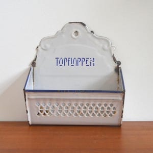 Könnte beinhalten: Weißer emaillierter Wand-Küchenbehälter mit dekorativer Oberseite und perforierter Front. Der Behälter hat blaue Verzierungen und das Wort "TOPFLAPPEN" in blauen Buchstaben. Der Behälter steht auf einer Holzoberfläche.