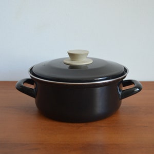 Könnte beinhalten: Schwarzer Emaille-Topf mit einem weißen Knopf auf dem Deckel. Der Topf steht auf einer Holzoberfläche.