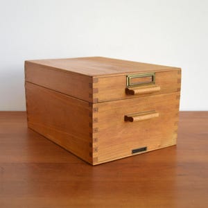Könnte beinhalten: Eine hellbraune Aufbewahrungsbox aus Holz mit zwei übereinanderliegenden Schubladen. Die obere Schublade verfügt über einen Messing-Etikettenhalter und einen Holzgriff, während die untere Schublade einen passenden Holzgriff besitzt.