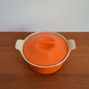 Könnte beinhalten: Orangefarbene Keramik-Auflaufform mit weißem Deckel und zwei Griffen. Die Form steht auf einer Holzoberfläche.