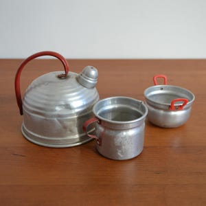 Könnte beinhalten: Ein Set aus drei Miniatur-Küchenutensilien aus Aluminium, bestehend aus einer Teekanne, einem Topf und einer kleinen Tasse. Alle drei Artikel haben rote Kunststoffgriffe.