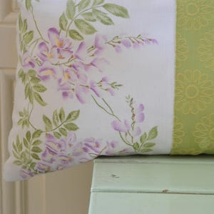 Könnte beinhalten: Ein dekoratives Kissen mit floralem Design. Eine Seite zeigt einen weißen Hintergrund mit lila Blauregen und grünen Blättern. Die andere Seite hat einen grünen Hintergrund mit einem gelben Blumenmuster. Das Kissen liegt auf einer hellgrünen Oberfläche.