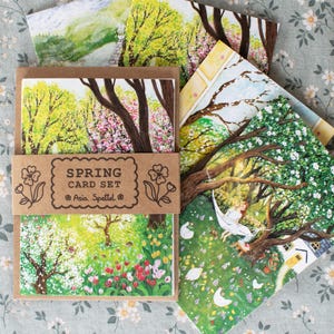 Peut inclure: Un ensemble de cartes de printemps avec des illustrations à l'aquarelle d'arbres et de jardins en fleurs. L'ensemble comprend plusieurs cartes avec des scènes différentes, liées par une bande de papier brun avec l'inscription "SPRING CARD SET" et le nom de l'artiste.