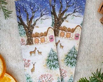 May include: Two rectangular winter scene bookmarks. Each bookmark features a snow-covered cottage, deer, and trees under a blue sky with falling snow. The bookmarks are surrounded by pine needles and dried orange slices.