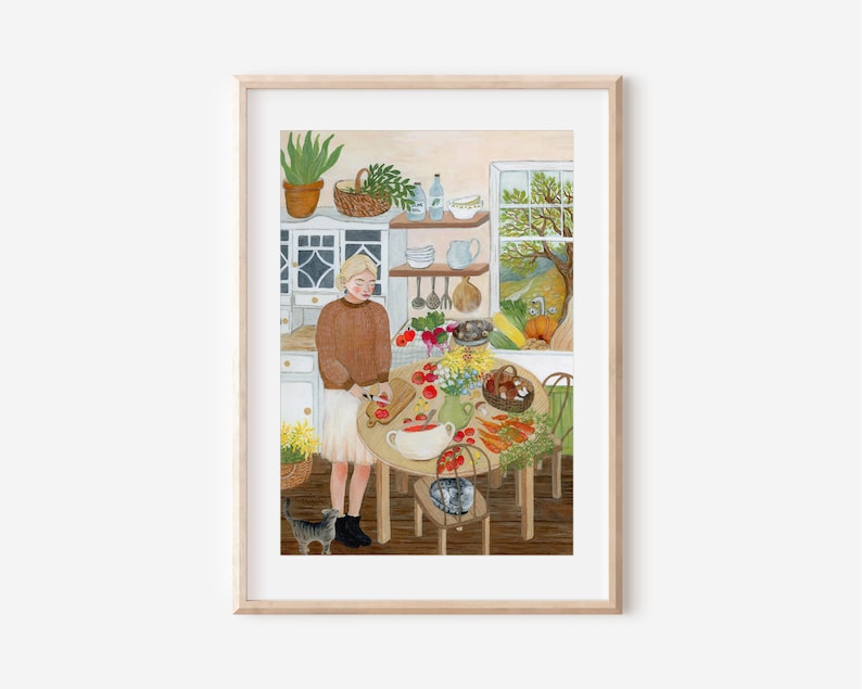 Op de afbeelding: Een ingelijste illustratie van een keukensc&egrave;ne met een persoon in een bruine trui en witte rok die tomaten snijdt op een houten plank aan een ronde tafel. Op de tafel staan groenten, een vaas met bloemen en een kom. Een kat staat op de vloer.