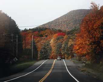 May include: A winding asphalt road with yellow lane markings leads through a forest of colorful autumn foliage. The trees are a mix of red, orange, yellow, and green. A single car drives down the road.