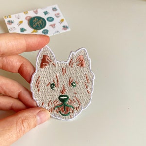 May include: White embroidered patch of a dog's face with pink ears, green eyes, and a brown nose. The dog is smiling with its tongue sticking out.