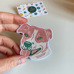 May include: Embroidered iron-on patch featuring a pink and white dog with green eyes. The dog is smiling with its tongue sticking out.