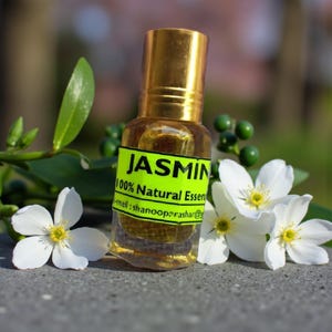Op de afbeelding: Een kleine glazen fles met een gouden dop, gevuld met een gouden vloeistof, met het label "JASMIN 100% Natural Essence". De fles is omgeven door witte bloemen met gele centra en groene bladeren.