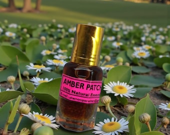 May include: A small amber-colored glass bottle with a gold cap, labeled "AMBER PATCH 100% Natural Essence." The bottle is surrounded by green leaves and white daisies with yellow centers, set against a blurred natural background.