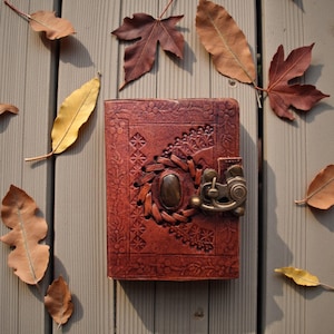 Puede incluir: Un diario de cuero marrón con intrincados patrones florales y geométricos en relieve. El diario presenta un diseño tejido alrededor de una piedra ovalada central y un cierre de metal decorativo. Hojas de otoño rodean el diario.