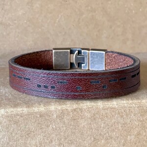 May include: A brown leather bracelet with a silver and brass clasp. The bracelet has a decorative pattern of lines and dots.