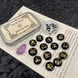 May include: A set of black and gold witch runes in a metal tin with the words "Witches Runes" and "Divination Arts" on the lid. The runes are on a white surface with text about casting runes.