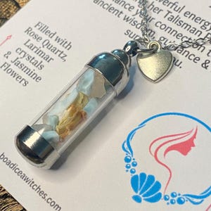 May include: A clear glass vial pendant filled with Rose Quartz, Larimar crystals, and Jasmine flowers. The pendant has silver-colored metal caps and a small heart charm. The necklace is on a silver chain.
