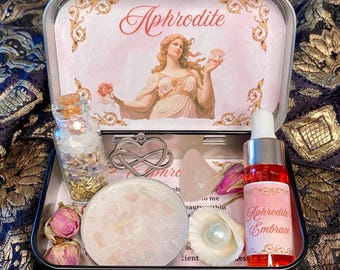 May include: A metal tin containing a small glass bottle with dried flowers, a silver heart-shaped infinity charm, a round candle with pink stones, a rose quartz crystal, two dried rosebuds, a white seashell with a pearl, and a red oil bottle labeled Aphrodite Embrace.