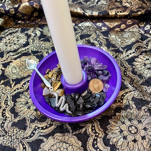 May include: A circular purple candle holder with a white candle. The holder is filled with crystals, a small silver spoon, and a coin. The crystals are in shades of brown, black, and purple. A silver snake charm is also present.