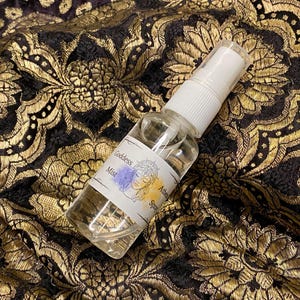May include: A small clear glass bottle with a white spray top. The bottle has a white label with the text "Goddess Mist" and a drawing of a woman's face with a crown. The label also has the text "Athena Spr"