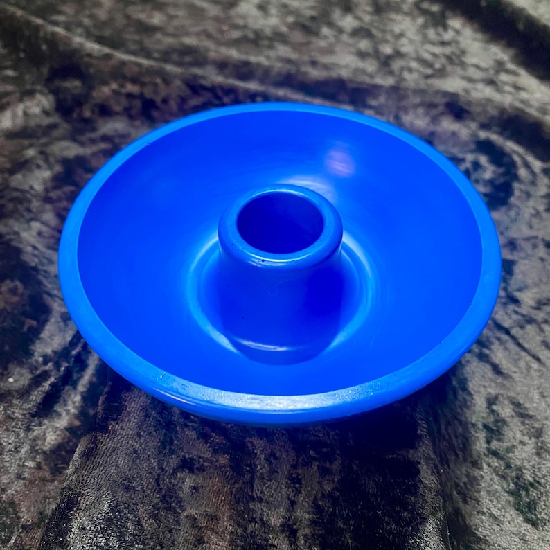 May include: A bright blue candle holder with a circular base and a central, raised cylinder to hold a candle. The holder is made of a smooth, solid material and is designed to catch any wax drips. The overall design is simple and modern.