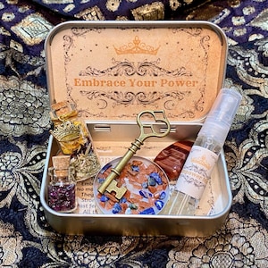 May include: A rectangular metal tin containing a small glass bottle with a cork and gold crown charm, a second glass bottle with purple stones, a brass-coloured key, a round container with red and blue stones, and a clear spray bottle labelled Empower Mist.