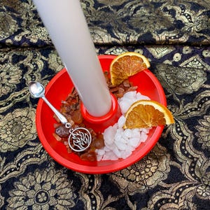 May include: A red candle holder with a white candle, dried orange slices, and crystals. A small silver spoon and a pendant with a flame design are also present. The background is a black and gold floral pattern.