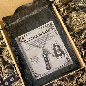 May include: A Goddess Hekate pendant necklace in a black mesh bag, inside a cardboard box. The necklace features a small glass vial filled with black onyx, lapis lazuli crystals, and cinnamon. The box also contains a card with text and an illustration.