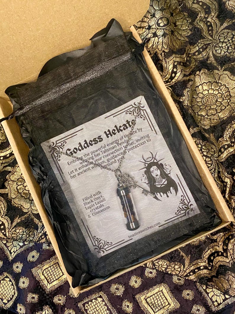 May include: A Goddess Hekate pendant necklace in a black mesh bag, inside a cardboard box. The necklace features a small glass vial filled with black onyx, lapis lazuli crystals, and cinnamon. The box also contains a card with text and an illustration.