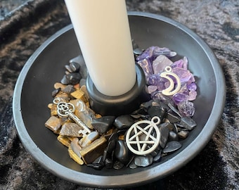 May include: A black candle holder with a white candle. The holder is filled with black, gold, and purple stones, and silver charms including a key, a crescent moon, and a pentagram. The candle holder is on a dark surface.
