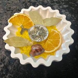 May include: A white, scalloped-edge dish filled with yellow rice, dried orange slices, bay leaves, and a small candle. A polished, brown and red stone and a decorative metal charm are also included. The arrangement is set against a dark background.