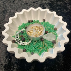 May include: A white, scalloped-edge dish contains green rice, a silver crescent moon charm, a small candle, and several dark berries. A clear, teardrop-shaped stone is also present. The dish is on a black surface.
