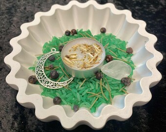Puede incluir: Un plato blanco con borde festoneado contiene arroz verde, un dije de media luna plateada, una vela pequeña y varias bayas oscuras. También hay una piedra transparente en forma de lágrima. El plato está sobre una superficie negra.
