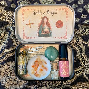 May include: A Goddess Brigid themed kit in a metal tin. The tin contains a candle with orange crystals, a green stone, a bottle of oil, and small bottles with herbs. The tin lid features an illustration of a woman and the text "Goddess Brigid".