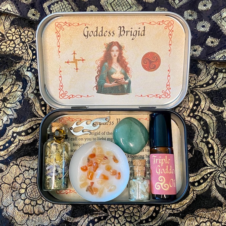 May include: A Goddess Brigid themed kit in a metal tin. The tin contains a candle with orange crystals, a green stone, a bottle of oil, and small bottles with herbs. The tin lid features an illustration of a woman and the text "Goddess Brigid".