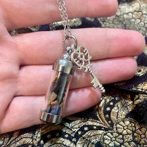 May include: Silver-toned necklace featuring a small glass vial filled with colorful stones and a decorative key charm. The vial has a silver cap and base. The key charm is ornate. The necklace is displayed against a patterned background.