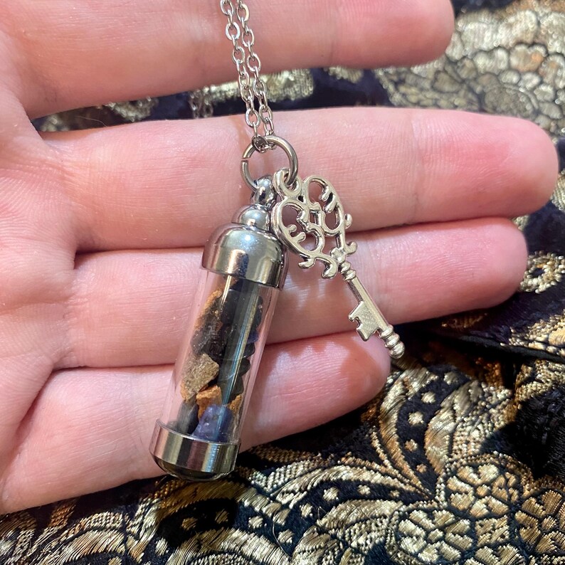 May include: Silver-toned necklace featuring a small glass vial filled with colorful stones and a decorative key charm. The vial has a silver cap and base. The key charm is ornate. The necklace is displayed against a patterned background.