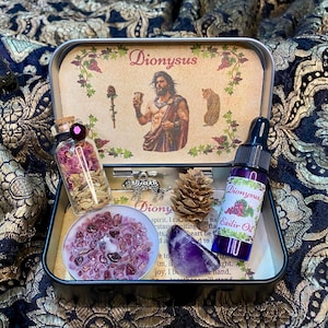 May include: A decorative metal box labelled "Dionysus", containing a small bottle of dried flowers, a candle, a crystal, a pinecone, and a bottle of "Exilir Oil". The box features an illustration of a man and a leopard.