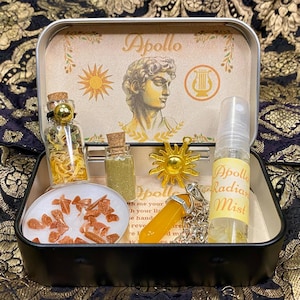 May include: A silver tin box with a gold and white design featuring a bust of Apollo, a sun, and a lyre. The box contains a variety of items, including a small bottle of dried herbs, a bottle of glitter, a candle, a sun charm, a crystal, and a bottle of mist labelled "Apollo Radiant Mist".