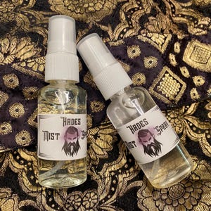 May include: Two clear spray bottles labelled "Hades Mist Spray" with a graphic of a bearded man. The bottles have white spray nozzles and are filled with a clear liquid. The background is a dark fabric with gold floral patterns.