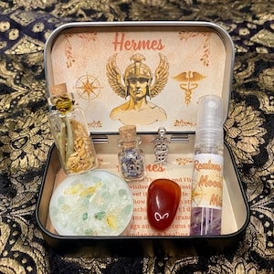 Op de afbeelding: Een decoratieve metalen doos met de naam "Hermes" op het deksel, met kleine glazen flesjes, een rode steen en een klein rond object met kristallen. De doos bevat ook een spuitfles met het label "Realm Moon Mist."
