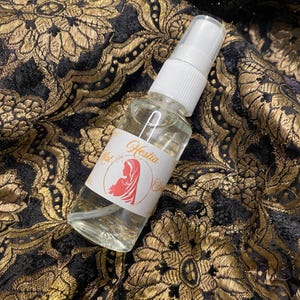 May include: A clear glass bottle with a white spray top and a white label that says "Hestia Mist" with a stylised image of a woman in a veil.