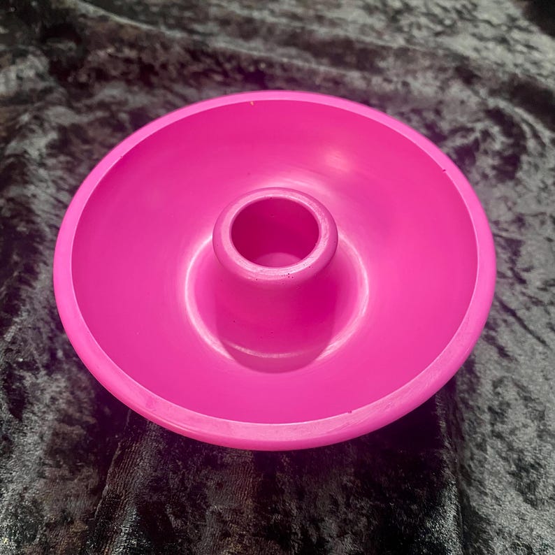 May include: A bright pink candle holder with a circular base and a central, raised cylinder for the candle. The holder is made of a smooth, solid material and sits on a dark, textured surface.