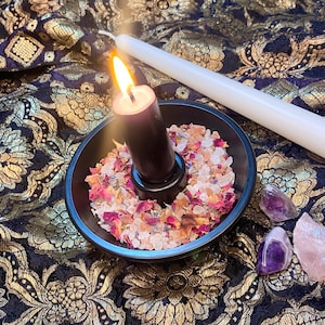 May include: A lit black candle in a black bowl filled with dried flower petals and crystals. A white candle lies next to the bowl on a dark fabric with gold floral patterns. Amethyst and rose quartz crystals are also present.