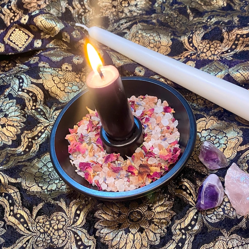 May include: A lit black candle in a black bowl filled with dried flower petals and crystals. A white candle lies next to the bowl on a dark fabric with gold floral patterns. Amethyst and rose quartz crystals are also present.