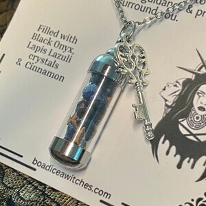May include: A silver-toned necklace featuring a clear vial pendant filled with blue crystals and cinnamon sticks. A decorative key charm hangs next to the vial. The text on the card reads "Filled with Black Onyx, Lapis Lazuli crystals & Cinnamon."