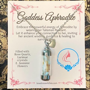 May include: A silver-toned necklace with a clear glass pendant filled with rose quartz, larimar crystals, and jasmine flowers. The card behind the necklace reads "Goddess Aphrodite" and includes text about the pendant's energy.